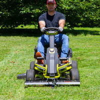 Wearing a red cap, black shirt, jeans, and gloves, a person expertly uses the Brinly Parts 38 in. Front Mount Dethatcher DT-38RY for the RYOBI Electric Riding Mower on lush grass. Scenic bushes and trees provide a picturesque backdrop to their lawn care routine.