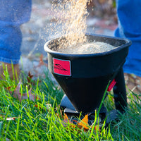 A person uses the Brinly Parts 5lb Capacity Handheld Spreader (HHS3-5BH) to distribute seeds or fertilizer on a lawn, spreading fine, granular material over the grass as autumn leaves create a colorful carpet below.