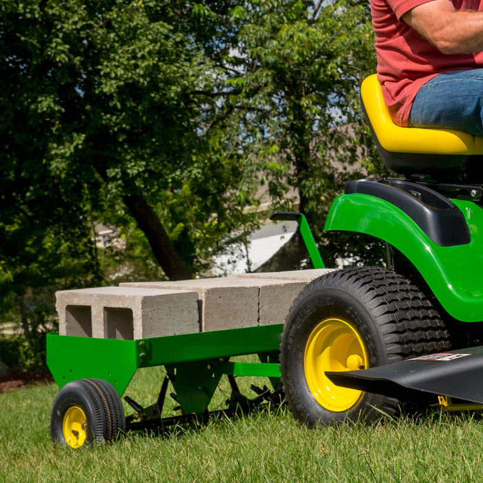 Wearing a red shirt, someone operates a John Deere 40 Tow-Behind Plug Aerator (PA-400JD/LPPA40JD) on a green ride-on lawn mower, pulling a trailer with gray concrete blocks. Vibrant trees and grass create the backdrop for aeration, promoting a healthy lawn under the clear sky.