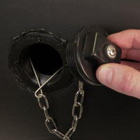 A hand holds a Brinly Parts 1019524 Tethered Plug, attached by a metal chain, beside an open circular port on a black surface—likely part of a tank or container.