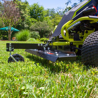 A close-up of a black Brinly Parts 38 in. Front Mount Dethatcher (DT-38RY) on a bright yellow RYOBI Electric Riding Mower shows its metal blade and wheel cutting grass. In the background, a garden with green trees and a teal umbrella appears under a clear blue sky, ideal for efficient lawn care.