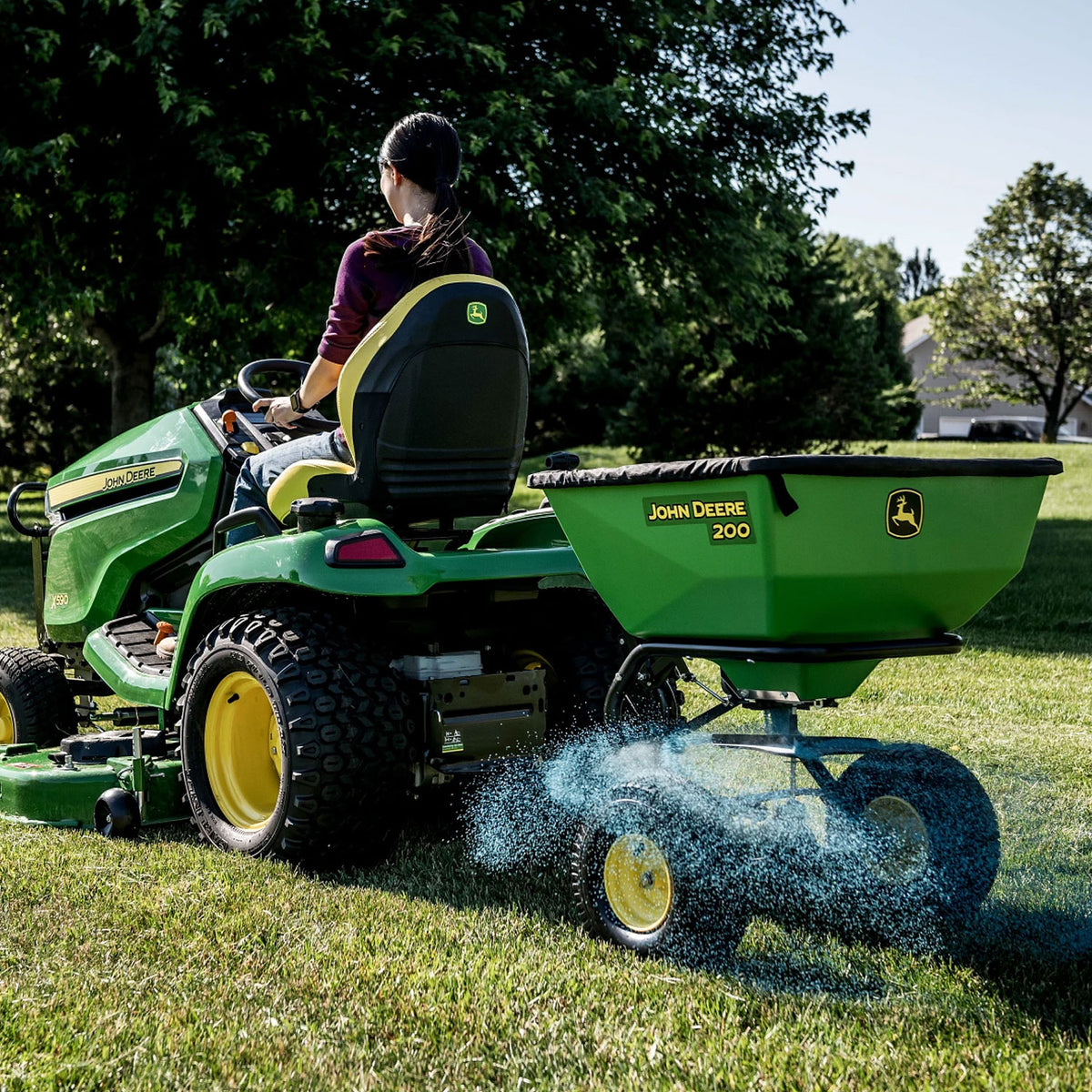 A person operates a John Deere 200 lb Tow-Behind Broadcast Spreader with ACCUWAY on a grassy lawn, demonstrating PRO-level durability. The Accu-Way™ technology ensures even distribution of blue fertilizer. Trees and a house are visible against a clear sky backdrop.