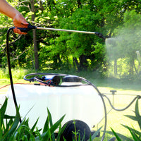 Using Brinly Parts ST-251BH 25 Gallon Tow-Behind Sprayer with Ultra Lo-Drift™ Spray Tips, a person efficiently distributes liquid fertilizer from a wheeled cart, promoting thriving green foliage and grass amid lush trees.