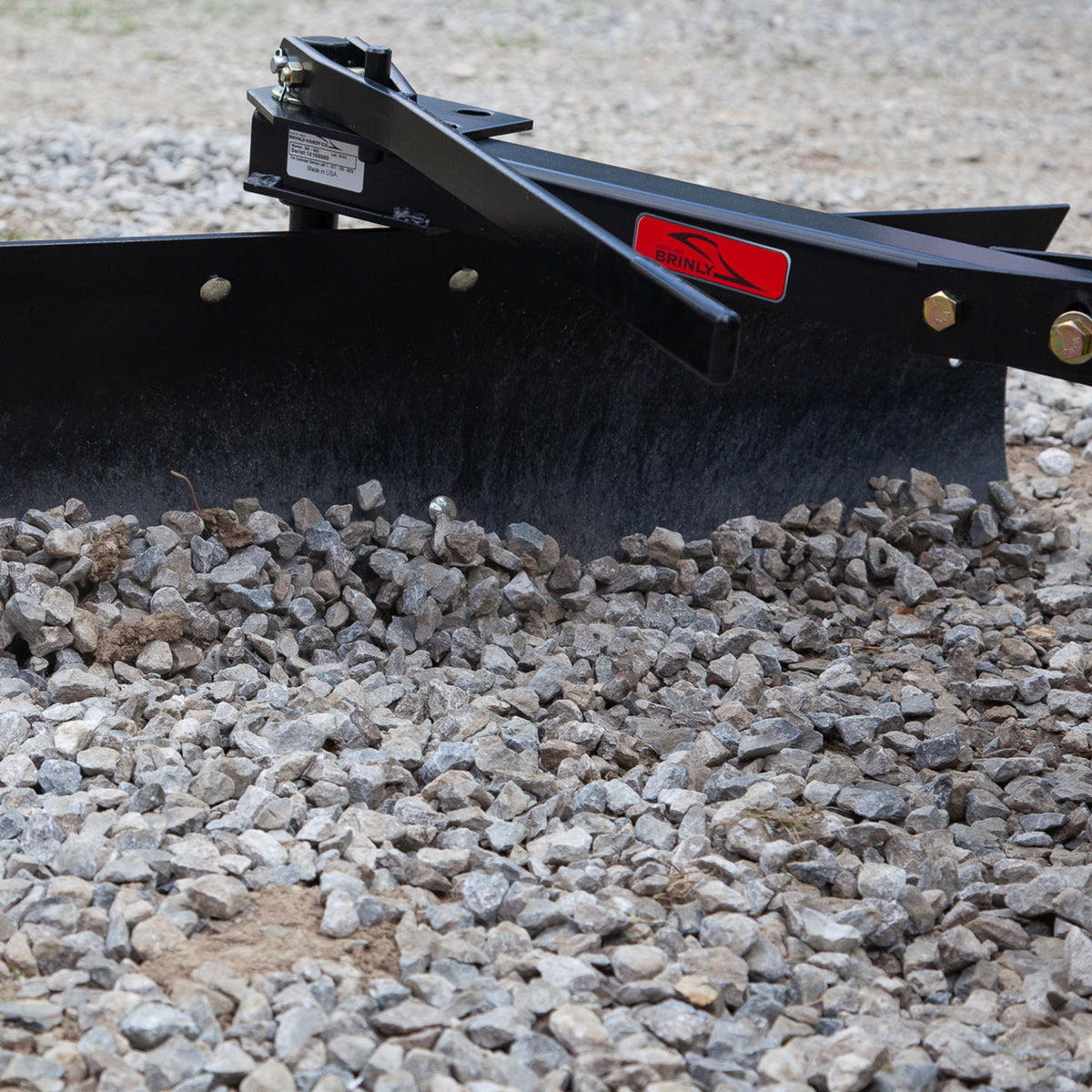 A close-up shows Brinly Parts 42 Sleeve Hitch Rear Blade | BB-562, an essential landscaping tool, attached to machinery. It smoothly grades a surface of gray gravel while the blurred background emphasizes the precision of this lawn tractor attachment.