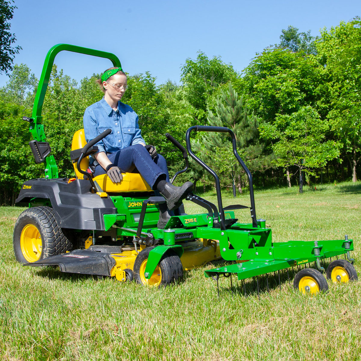 A person expertly drives a green John Deere Z-Trak, equipped with a Front-Mount 48 ZTR Dethatcher (CPLP83252), across a grassy area with trees. Clad in a blue denim shirt, gloves, and green headband, they skillfully manage the task on this sunny day.