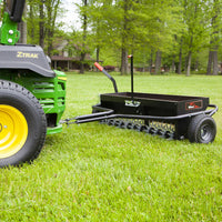 A Brinly Parts 40 Combination Aerator Spreader (AS2-40BH-G) with pneumatic tires, a black metal-weight tray, and a 100 lb. capacity is hitched to a lawn tractor on a green lawn. Trees and a house are in the sunny background.