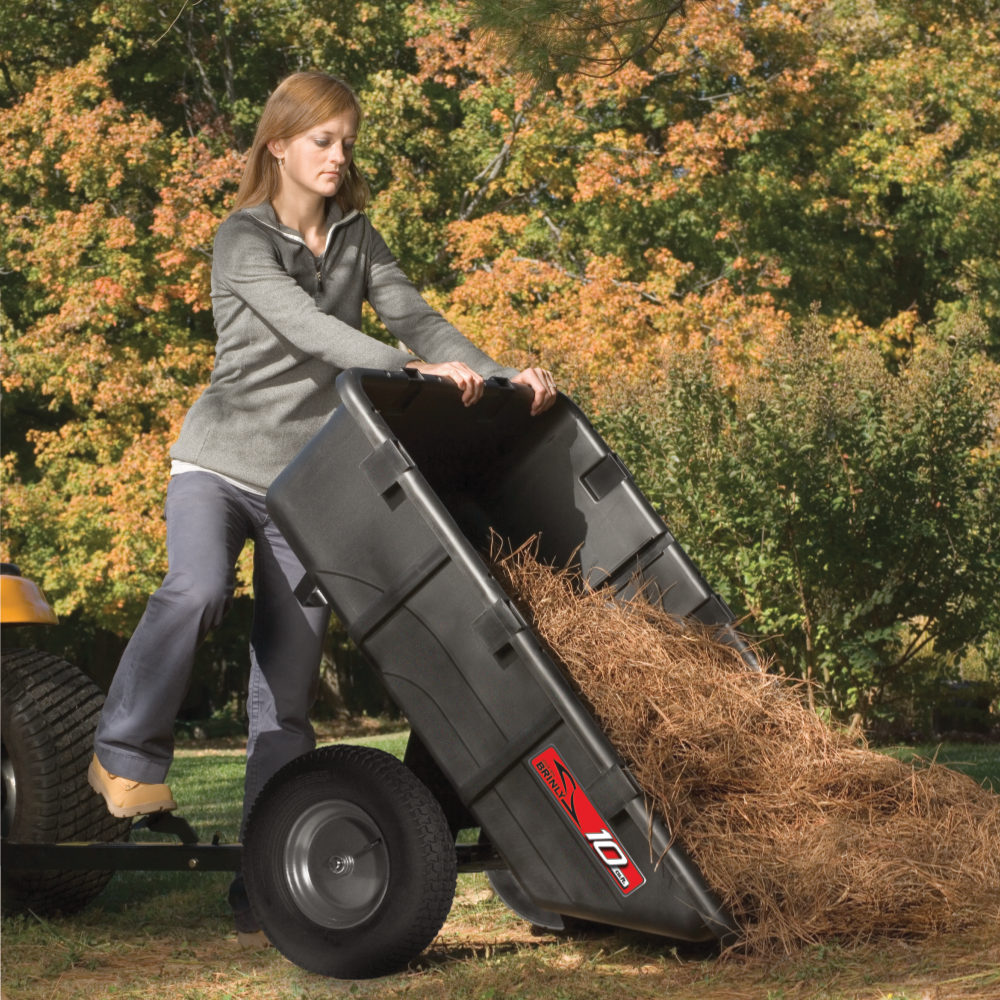 A woman in casual clothes empties a Brinly Parts 10 Cu. Ft. Poly Cart (PCT-101BH) filled with straw or pine needles onto the grass in a yard with autumn trees in the background.