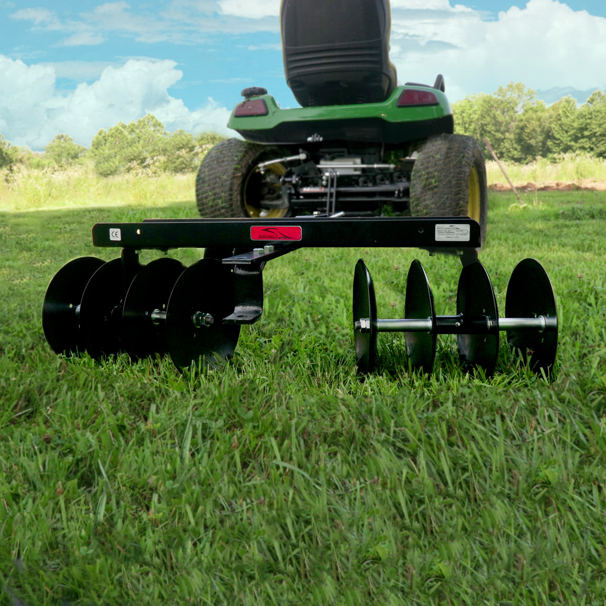 A green lawn tractor is parked on grass with a Brinly Parts Sleeve Hitch Disc Harrow | DD-551BH in the foreground, ideal for soil preparation. Multiple rotating disks are visible against a serene backdrop of trees and clouds.