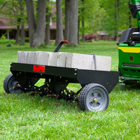 A Brinly Parts 48 Tow-Behind Plug Aerator (PA-482BH), featuring durable steel tines and concrete blocks, sits on a lawn. Connected to a green tractor, it efficiently penetrates the root zone. In the blurred background, trees and a house enhance the picturesque scene.