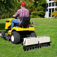 A person in a red cap and plaid shirt rides a yellow lawn tractor, towing the Brinly Parts Spike Aer 40 spike aerator weighted with concrete blocks for soil aeration on a grassy lawn near a house.