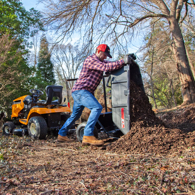 Wearing a red cap and plaid shirt, someone uses a garden tractor with The Roger 180-Degree Full Dump Kit for Brinly-Hardy 10 cu. ft. Poly Carts by Brinly Parts to efficiently dump mulch onto a garden area. Leafless trees are in the background, and the ground is covered with soil and leaves.