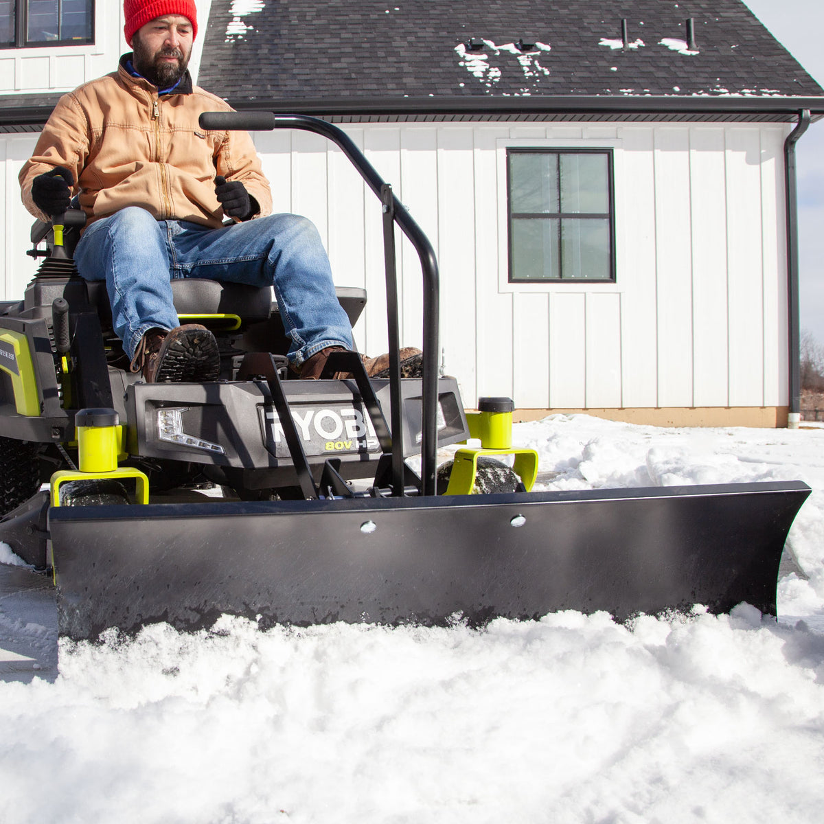 A person in a red beanie and brown jacket uses the Brinly Parts Front-Mount Blade for RYOBI Electric ZTRs (FB-42RYZT) to clear snow outside a white house with black trim, highlighting the steel blades powerful precision on this sunny, snowy day.