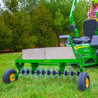 A John Deere 40 Spike Aerator with Rigid Steel Weight Tray, featuring galvanized steel stars and a space for concrete blocks, is attached to a tractor on grass. Its spiked wheels boost efficiency, set against a backdrop of evergreen trees.