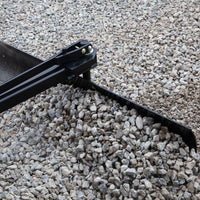 The Brinly Parts 42” Sleeve Hitch Rear Blade (BB-562), a versatile landscaping tool, evenly spreads a pile of gray rocks on a rough surface. Its black metal blade pushes the stones, creating a uniform gravel layer.