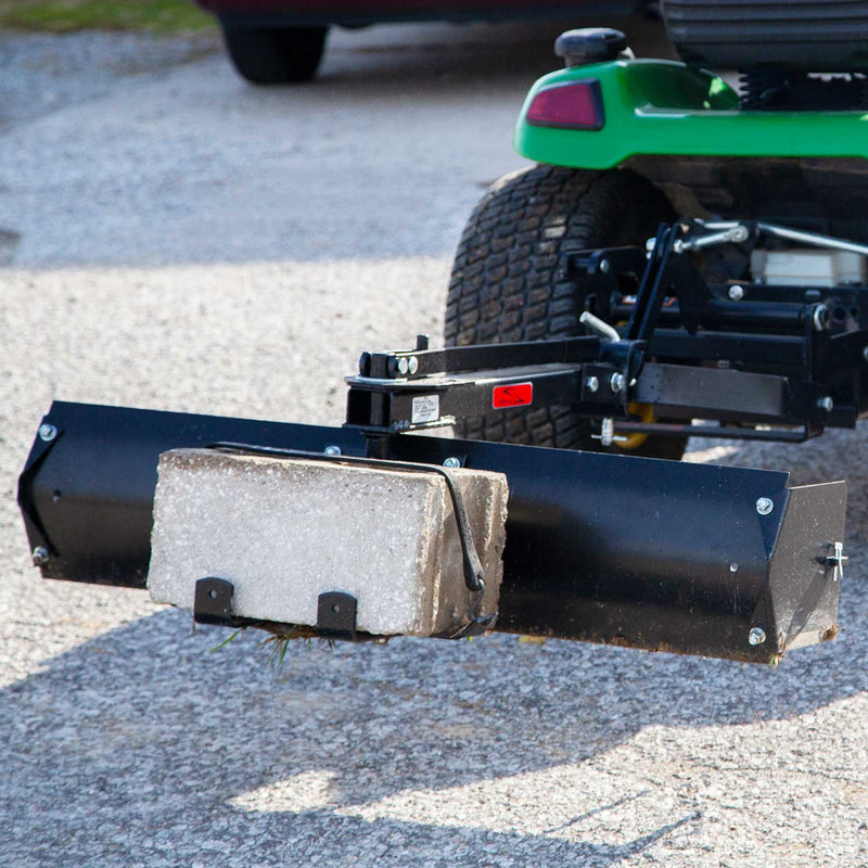 A green lawn tractor has a black Brinly Parts 42” Sleeve Hitch Box Scraper (BS-42BH) with a durable carbon steel blade for leveling. It includes a large cinder block for added weight, positioned on a gravel path with a parked vehicle visible in the background.