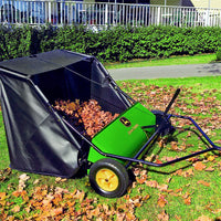 The green and black John Deere 42 in. 24 cu. ft. Tow-Behind Lawn Sweeper, equipped with two large yellow wheels, is filled with dry leaves and rests near a hedge and white fence on the lawn.