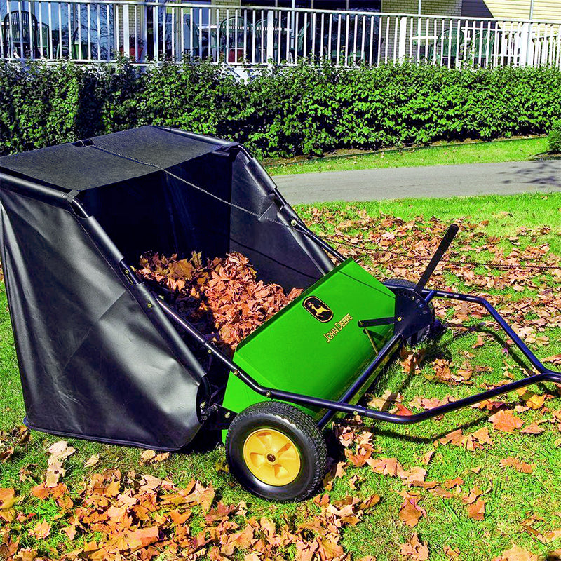 The green and black John Deere 42 in. 24 cu. ft. Tow-Behind Lawn Sweeper, equipped with two large yellow wheels, is filled with dry leaves and rests near a hedge and white fence on the lawn.