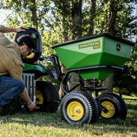 Wearing safety glasses, a person attaches a green John Deere Tow-Behind Broadcast Spreader (Model: LP79931) to their ride-on lawnmower outdoors on grass, with trees creating a serene backdrop.