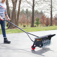 On a dry, slightly overcast day, someone in jeans and a gray T-shirt uses the 20 Push Spike Aerator with an adjustable handle by Brinly Parts on a paved path. The 3D galvanized steel tines ensure effective performance even in tough soil, with bare and evergreen trees visible in the background.