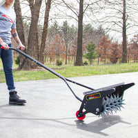 Wearing jeans, a gray t-shirt, and black boots, a person pushes a Brinly Parts 20 Push Spike Aerator with red wheels on a paved path. Featuring an adjustable handle and 3D galvanized steel tines, its perfect for yard care against a serene backdrop of trees and grass.