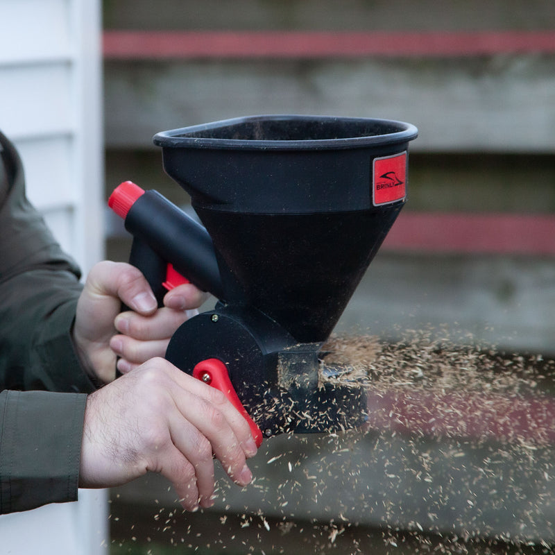 A person skillfully uses a Brinly Parts 5lb Capacity Handheld Spreader | HHS3-5BH to evenly disperse seeds across the lawn. The black device with its red handle and logo stands out against the white building and wooden fence backdrop.