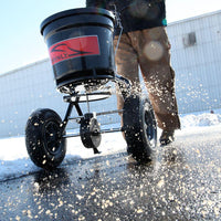 Using a Brinly Parts 50 lb. Push Spreader (P20-500BH) with a black container, a person in brown pants scatters salt on snowy pavement diligently, with a large building as the backdrop.