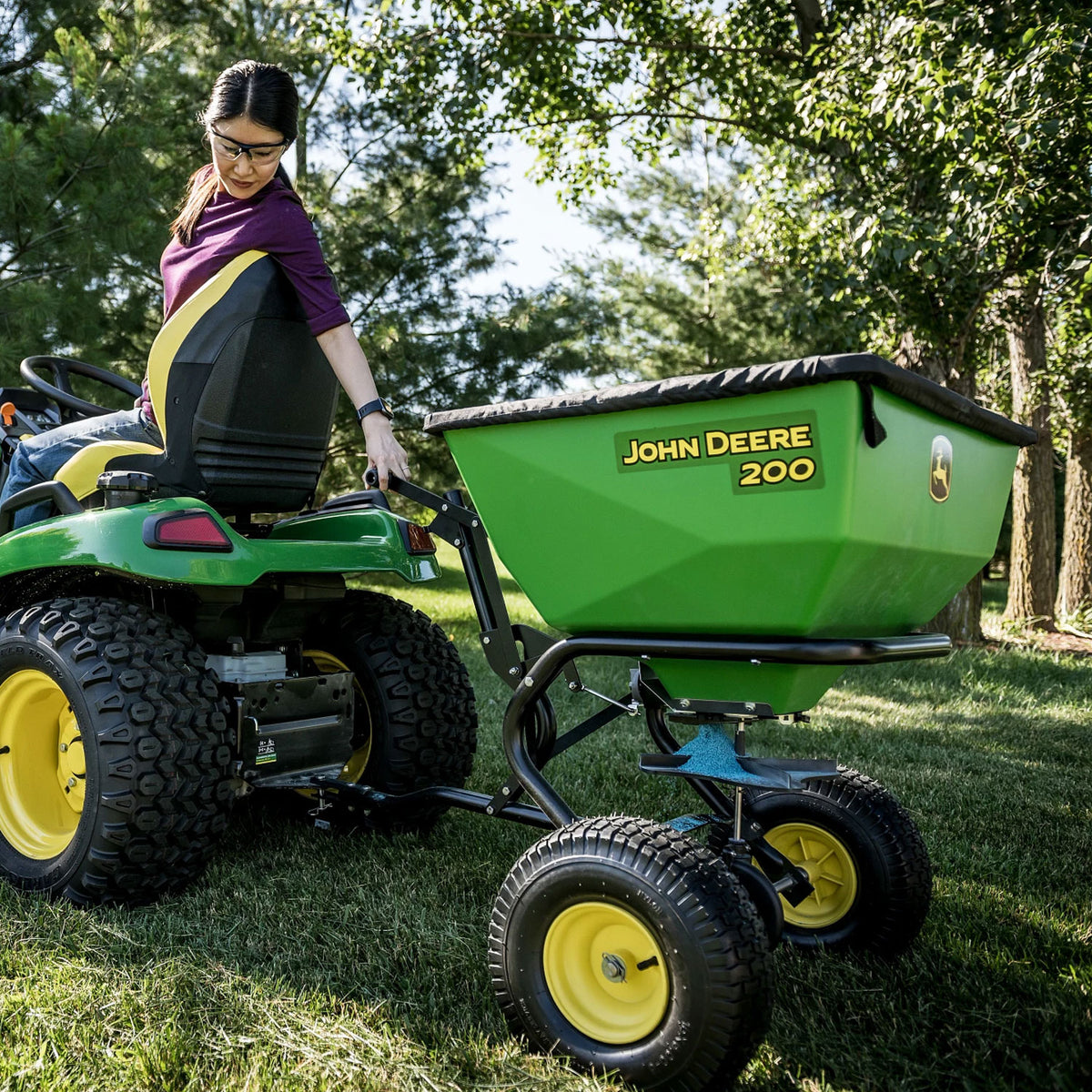 A dark-haired person in a purple shirt and glasses sits on a durable John Deere lawn tractor, attaching a green and black John Deere 200 lb Tow-Behind Broadcast Spreader with ACCUWAY | LP79932 trailer. The tranquil outdoor scene features grass and trees.