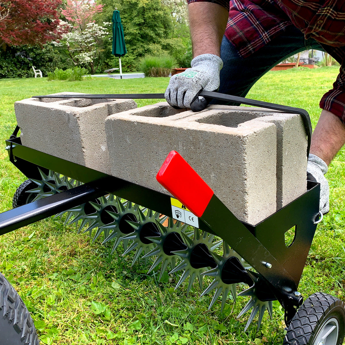 Wearing gloves, a person uses the Brinly Parts 40″ Tow-Behind Spike Aerator (SAT2-40BH-G) with spiked wheels and concrete block weights to aerate the lawn efficiently. In the background, a garden and umbrella enhance the serene setting.