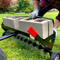 Wearing gloves, a person uses the Brinly Parts 40″ Tow-Behind Spike Aerator (SAT2-40BH-G) with spiked wheels and concrete block weights to aerate the lawn efficiently. In the background, a garden and umbrella enhance the serene setting.