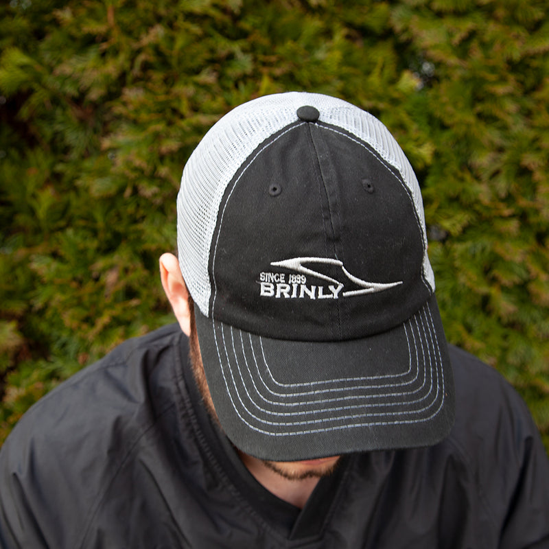 A person in a Brinly Parts Gray & Black Baseball Cap and dark jacket stands outside before green shrubs, their face mostly hidden as they look down.