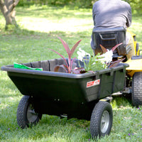 A person on a yellow lawn tractor pulls a Brinly Parts 10 Cu. Ft. Poly Cart (PCT-101BH) filled with potted plants, green gloves, and gardening tools across a grassy yard.