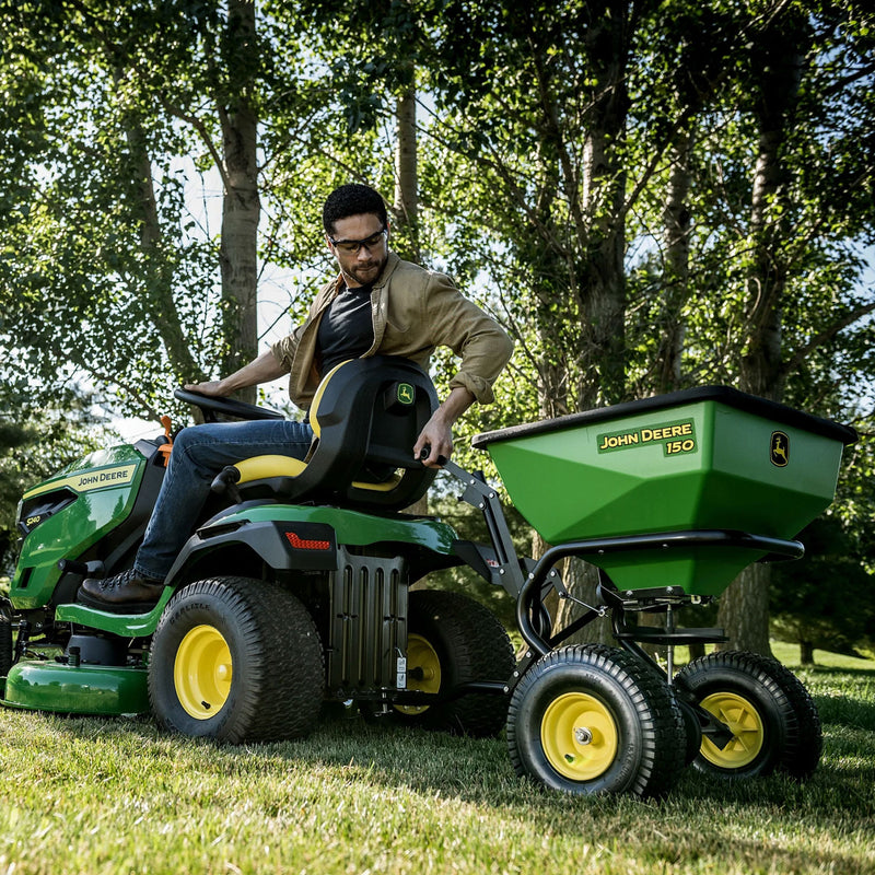 A glasses-wearing individual operates a John Deere riding mower in a grassy, tree-lined area, with sunlight creating dappled effects. The rugged durability of their 150 lb Tow-Behind Broadcast Spreader with ACCUWAY (LP79931) ensures smooth towing.