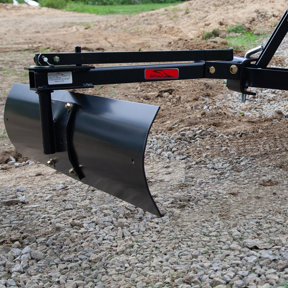 A close-up of the Brinly Parts 42” Sleeve Hitch Rear Blade | BB-562, a black metal attachment for landscaping or lawn tractors, over gravel and dirt.