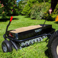 Someone is using the Brinly Parts 40 Combination Aerator Spreader with Pneumatic Tires (AS2-40BH-G) to tend green grass. The aerator features sturdy spiked wheels and a 100 lb. capacity hopper, ideal for soil enhancement, set against a serene backdrop of lush greenery and flowering bushes.