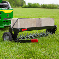 A Brinly Parts 40″ Spike Aerator with Transport Wheels & 3D Galvanized Steel Tines (SAT-401BH) is towed behind a green lawn tractor on grass, using concrete blocks for effective lawn aeration.