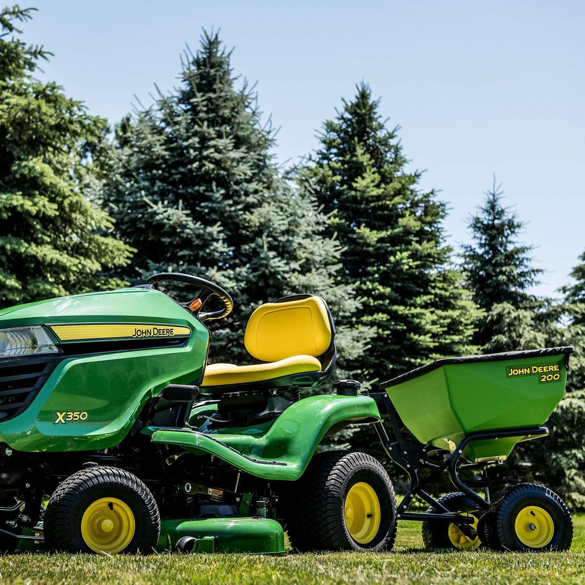 A John Deere X350 lawn tractor with PRO-level durability is paired with a John Deere 200 lb Tow-Behind Broadcast Spreader. Both rest on grass under tall evergreens and a clear blue sky.