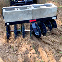 Close-up of the Brinly Parts Sleeve Hitch Disc Harrow | DD-551BH, featuring multiple rotating discs, partially submerged in dirt. Two concrete blocks on top add weight for deeper soil preparation against a backdrop of freshly tilled earth.