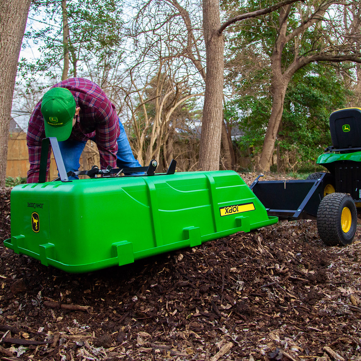 In a wooded area, someone in a plaid shirt and cap loads soil into a John Deere 180 Degree Full Dump Cart, showcasing its 650 lb. hauling capacity. The green tow-behind cart is attached to a small tractor among bare trees and brown foliage, ready for action.