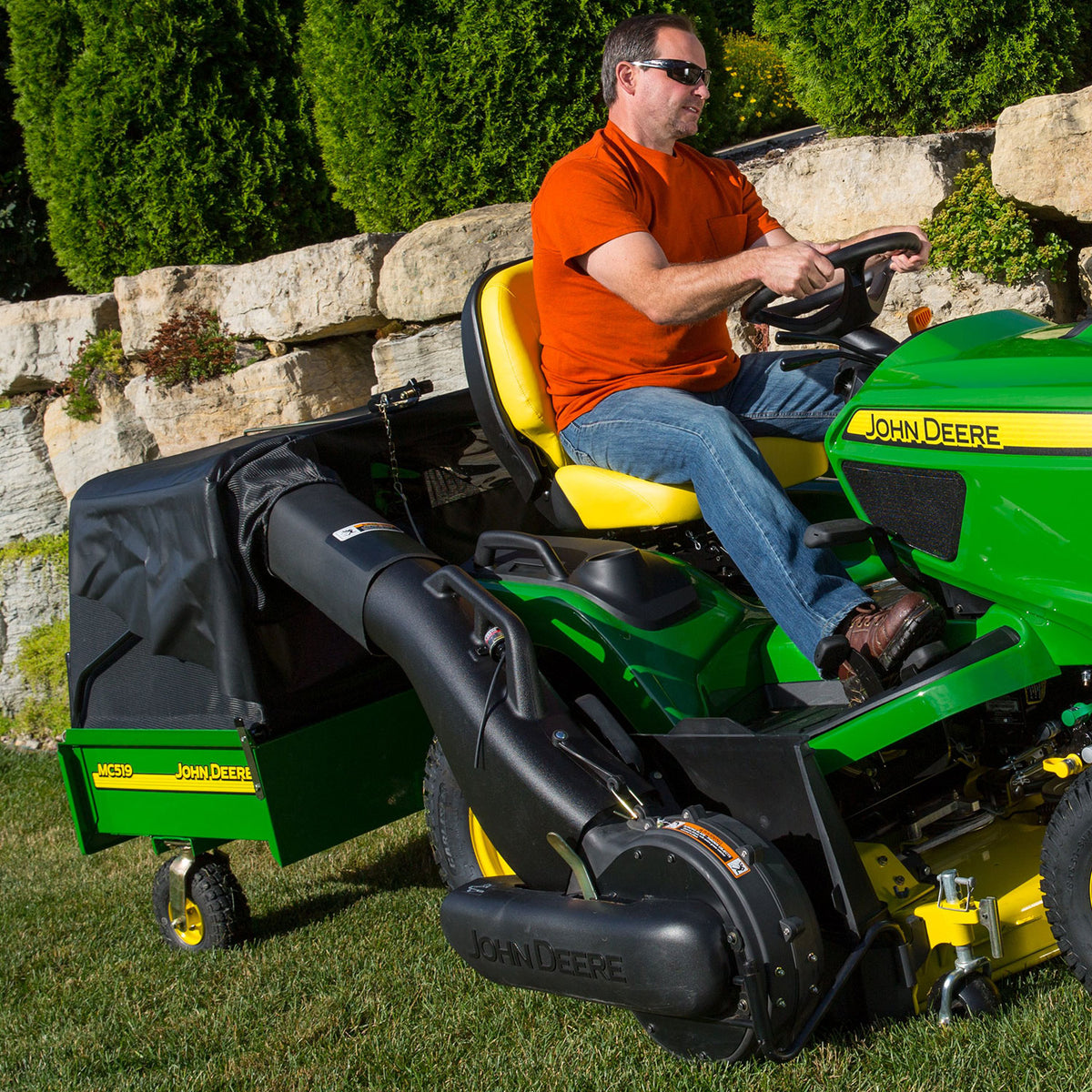 A man wearing sunglasses and an orange shirt rides a green John Deere mowing a grassy area bordered by stone walls and bushes, efficiently collecting clippings with the John Deere MC519 Material Collection System LP49228.