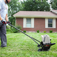A person flattens grass with a manual lawn roller in front of a brick house with white shutters, while the Brinly Parts 20 Push Spike Aerator with 3D Galvanized Steel Tines is nearby for effective soil aeration. Trees and shrubs are in the background.