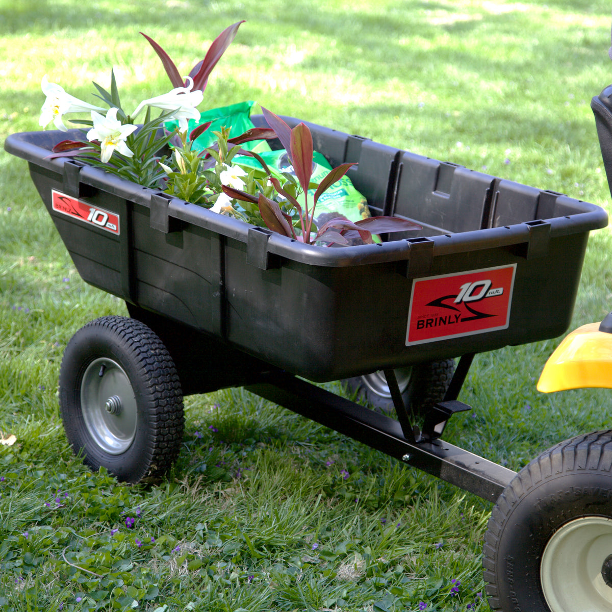 A Brinly Parts 10 Cu. Ft. Poly Cart | PCT-101BH is hitched to a yellow vehicle, transporting potted plants and gardening supplies across green grass outdoors.
