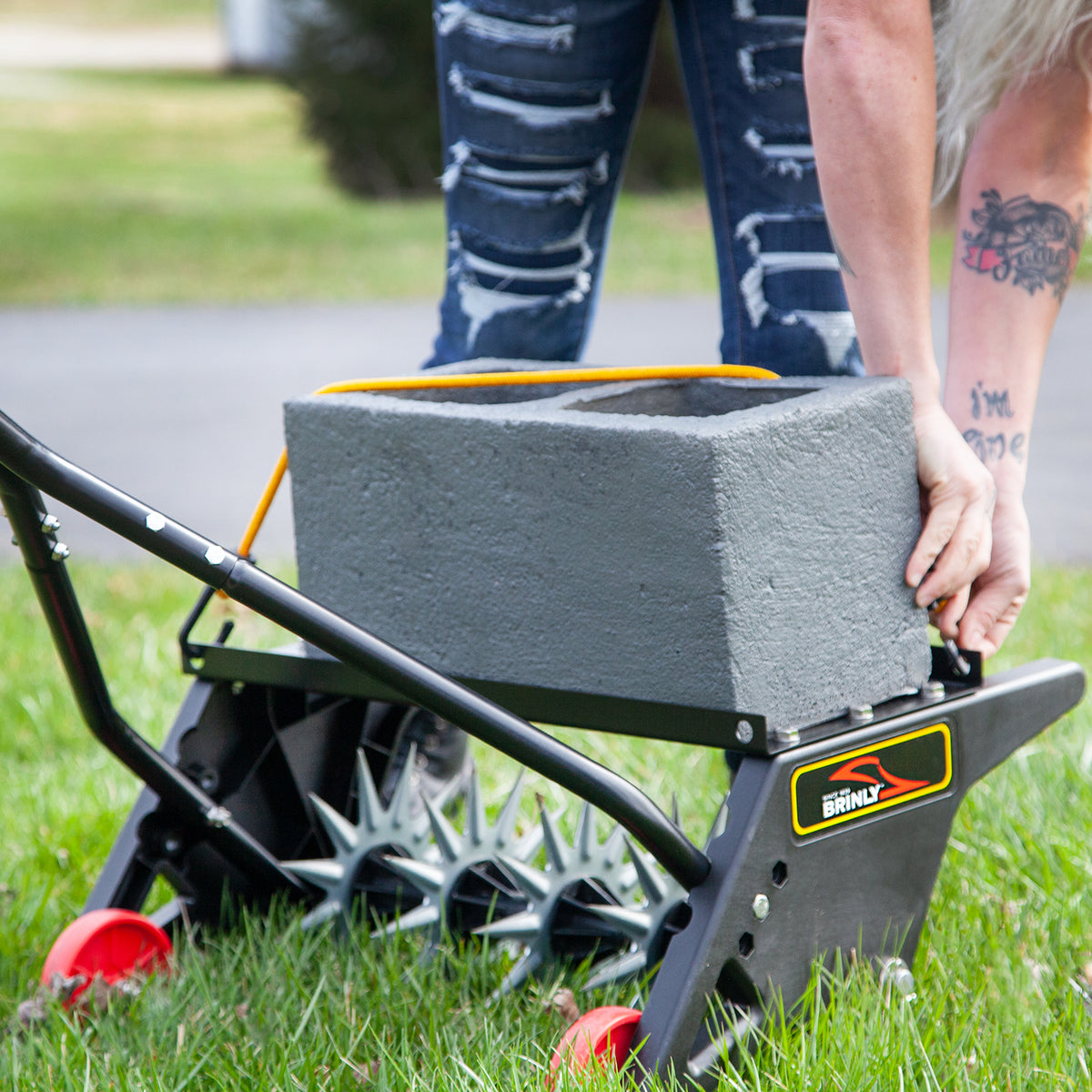 A person with tattooed arms and ripped jeans is placing a large gray concrete block onto a Brinly Parts 20 Push Spike Aerator, featuring 3D galvanized steel tines, resting on the grass.