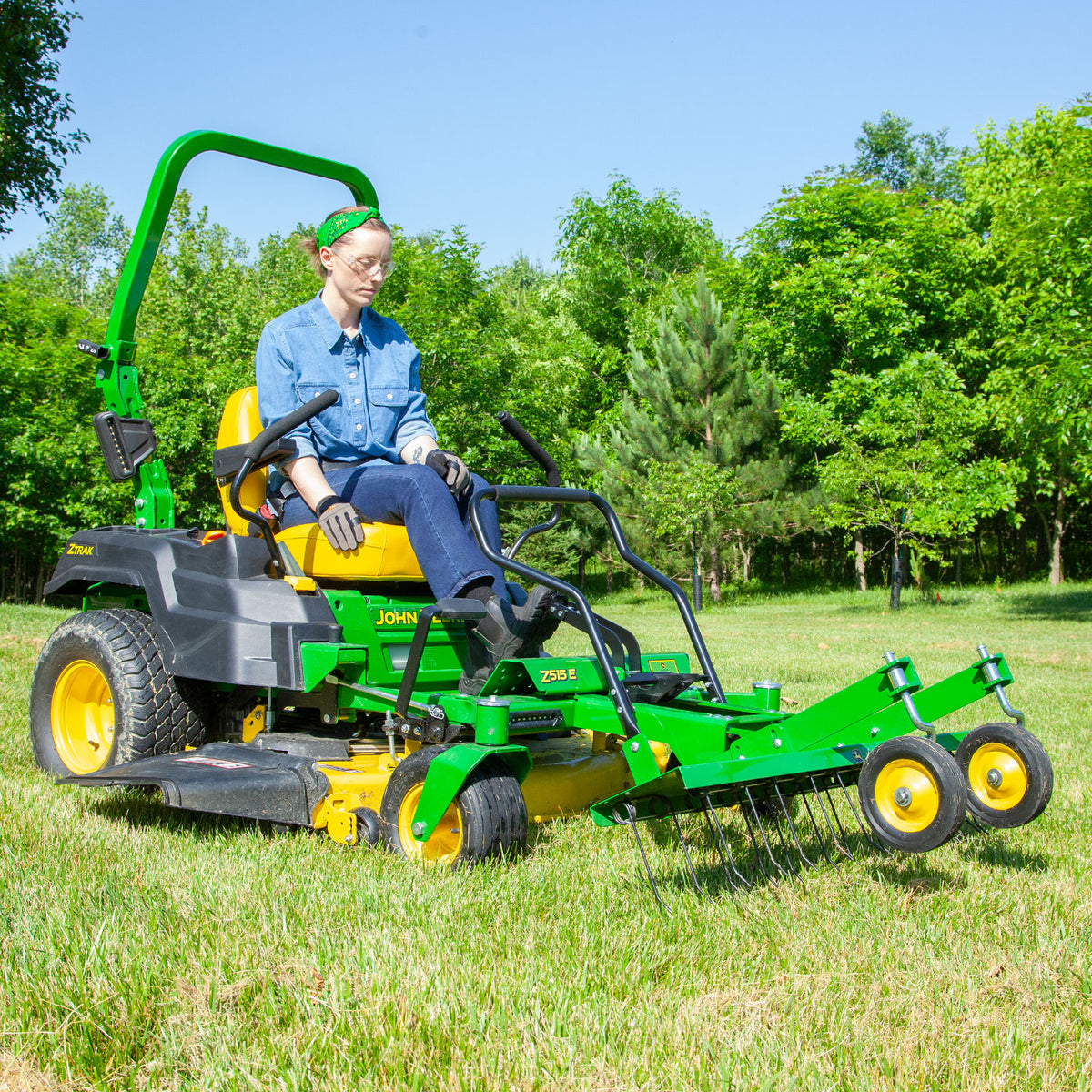 Wearing jeans and a denim shirt, a person skillfully maneuvers a John Deere Z-Trak Mower fitted with flexible spring steel tines and a John Deere Front-Mount 48 ZTR Dethatcher (CPLP83252) across a grassy field, framed by trees under a clear blue sky.