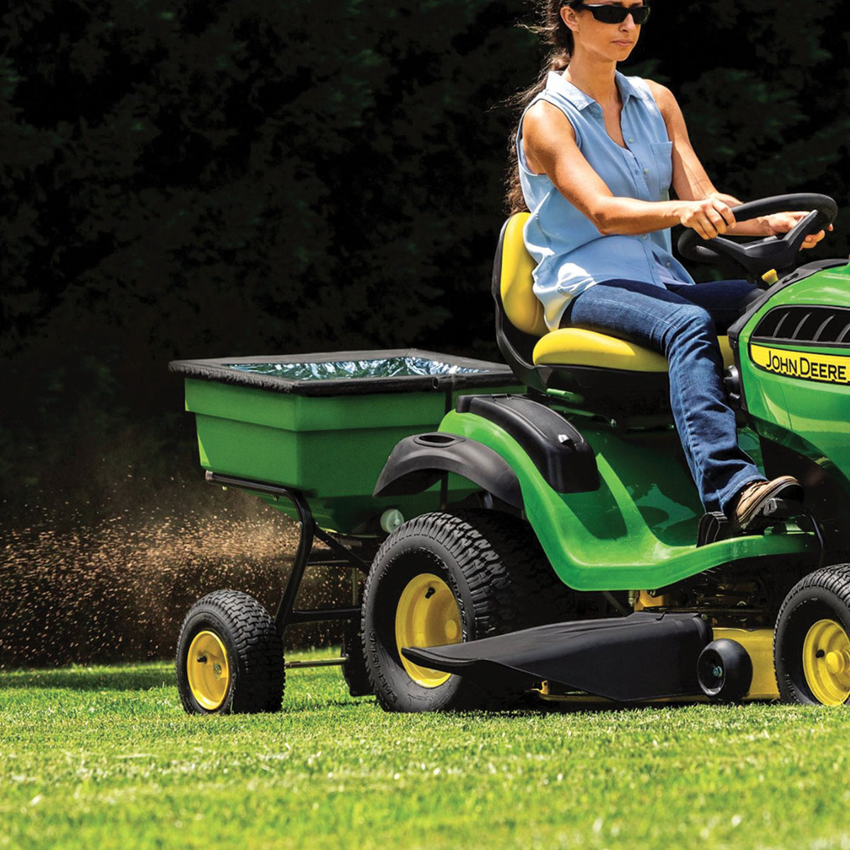 A person in sunglasses, jeans, and a light blue sleeveless top maneuvers a green John Deere Tow Broadcast Spreader - 125 lb across the sunlit lawn. The well-maintained grass and surrounding trees reflect the precision and quality of their equipment.