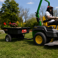 A person drives a green riding lawn mower pulling a Brinly Parts 10 Cu. Ft. Poly Cart (PCT-101BH) filled with colorful flowers on a sunny day, with trees and bushes in the background.
