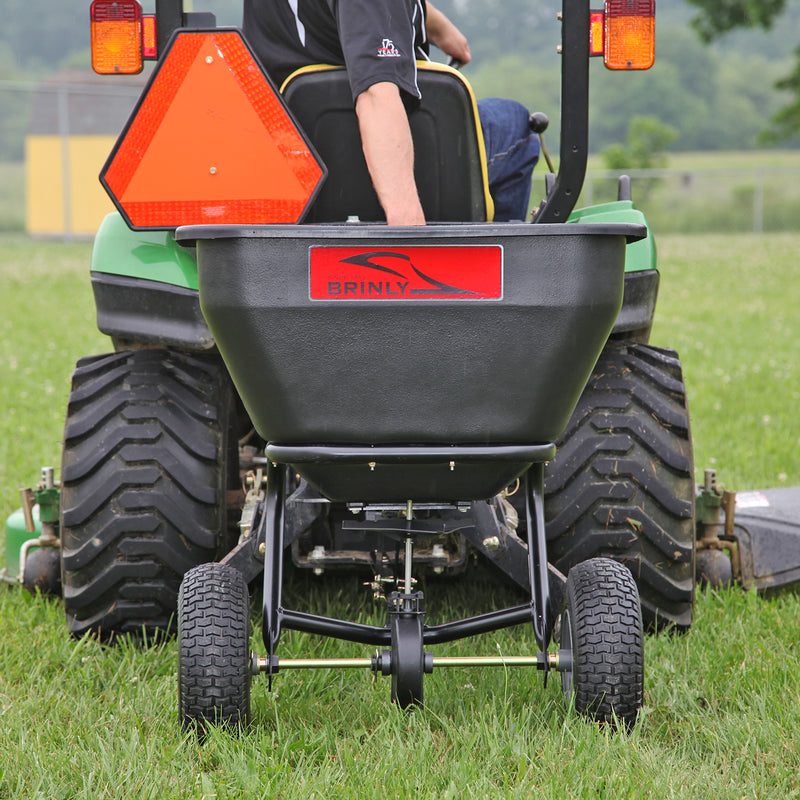 A person drives a green lawn tractor with a mounted black Brinly 175 Lb. Tow-Behind Spreader, BS36BH model, on a grassy field. The rust-proof polyethylene spreader has large tires and an orange triangle sign, promising durability and efficiency.