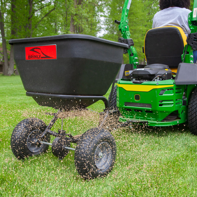 A person uses a Brinly Parts 175 Lb. Tow-Behind Spreader | BS36BH with their green lawn tractor to evenly distribute seeds over a lush lawn, set amid trees and greenery indicative of spring or summer.