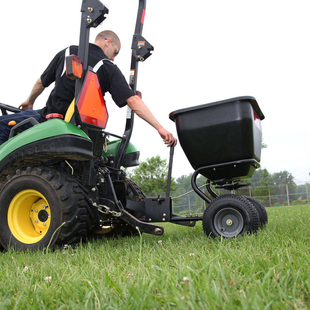 A person wearing a black shirt operates a green tractor with a black trailer on a grassy field, adjusting the lever of the Brinly Parts 175 Lb. Tow-Behind Spreader | BS36BH. The scene is framed by trees under an overcast sky.