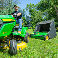 On a sunny day, someone uses a John Deere lawnmower with the 42 in. 24 cu. ft. Tow-Behind Lawn Sweeper to collect grass on a lush, green lawn bordered by trees and a house. The setup highlights visible branding and includes a yellow deck for efficient operation.