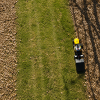 A yellow lawn mower is trimming grass, creating a distinct line between the lush green lawn and a brown, leaf-covered area as seen from above—the ideal scenario to showcase efficient leaf removal with the Brinly Parts 42″ Lawn Sweeper with DOUBLE-HELIX Brushes (LS2-42BH2-G), easily towed behind your mower.